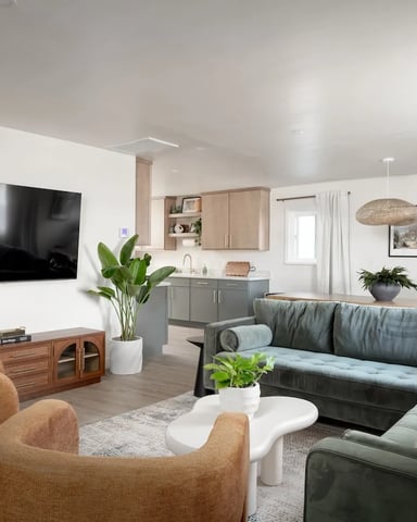 Open-plan living room with wall-mounted TV, gray sectional sofa, tan armchairs, wood media console, and kitchen with gray cabinetry visible in background.
