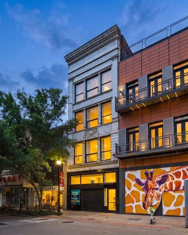 Urban street at dusk with historic and modern mixed-use buildings, ground-floor retail, and lit windows