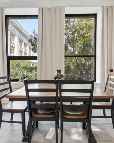 Bright dining room with black table, four chairs, cream curtains, and three large windows overlooking city buildings and trees