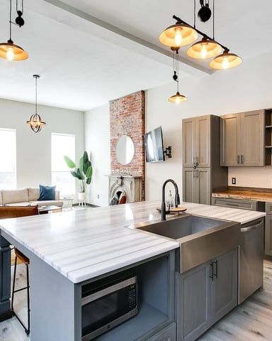 Open-plan kitchen and living room with marble island, stainless steel appliances, exposed brick wall, and pendant lighting.