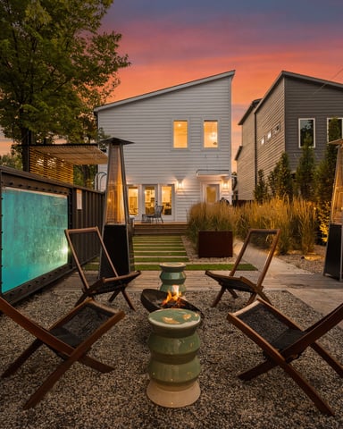 Modern vacation rental with backyard fire pit, lounge chairs, and glowing windows at dusk under colorful sunset sky