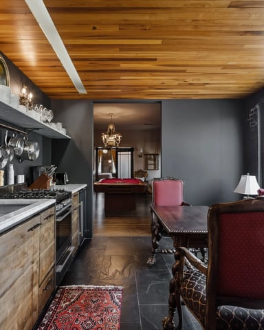 Open-plan kitchen and living area with wood ceiling, dark walls, stainless steel appliances, and burgundy seating overlooking city street.