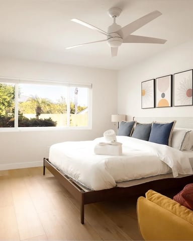 Bright bedroom with queen bed, ceiling fan, large windows overlooking greenery, desk, and yellow armchair