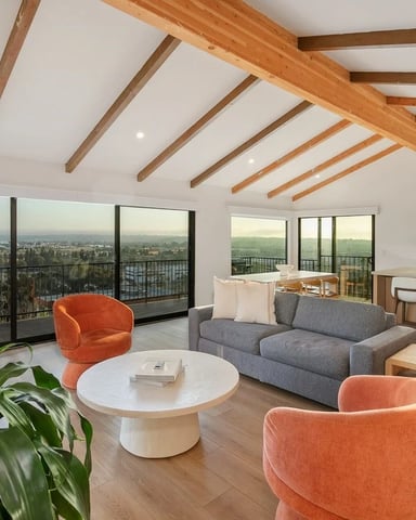 Modern open-plan living room with exposed wood beams, gray sofa, orange chairs, and expansive city views through floor-to-ceiling windows.