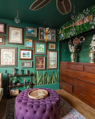 Eclectic bedroom with deep green walls, cream leather bed, purple tufted ottoman, wooden dresser, gallery wall of framed artwork, hanging plants and woven