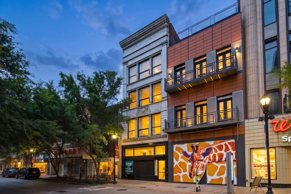 Urban street at dusk with historic and modern mixed-use buildings, ground-floor retail, lit windows, and street lamps