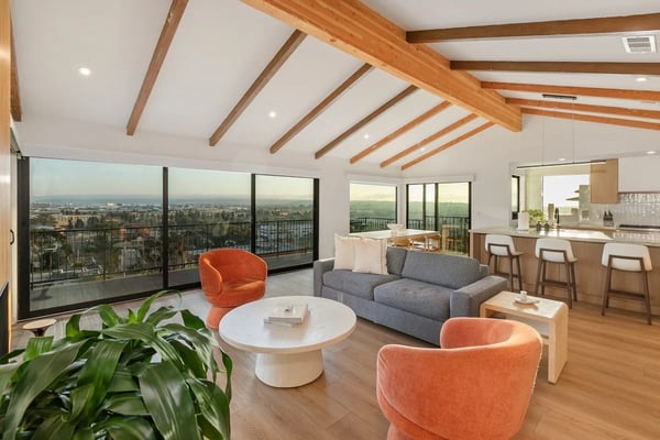 Modern open-plan living room with exposed wood beam ceiling, gray sofa, orange chairs, and expansive city views through floor-to-ceiling windows.