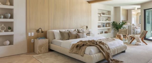 Modern bedroom with cream upholstered bed, wood paneled accent wall, built-in shelving, and open ensuite bathroom visible through glass door