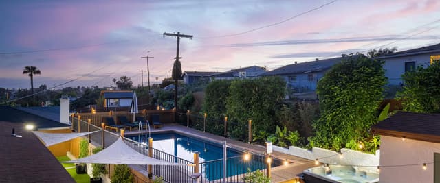 Backyard with illuminated pool, hot tub, and deck at dusk with mountain views and neighboring houses